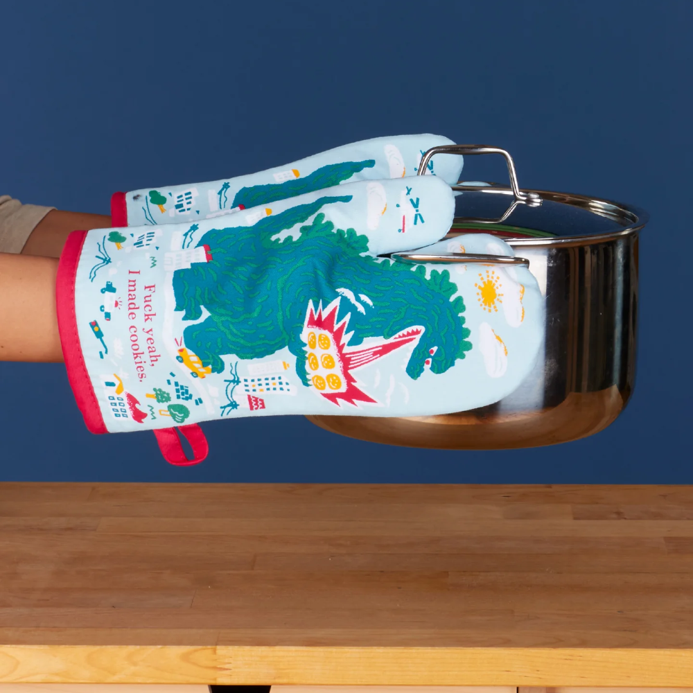 A person's hand wearing a colorful printed oven mitt while holding a stainless steel pot over a wooden countertop against a dark blue background.
