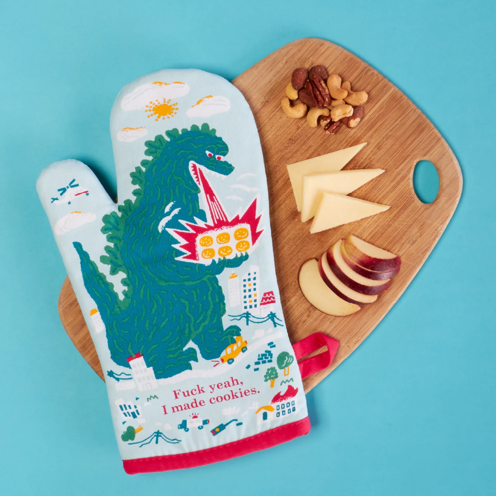 A novelty oven mitt featuring a green dinosaur riding a red hot air balloon with the text 'Fuck yeah, I made cookies.' styled on a light blue background next to a wooden cutting board with cheese and nuts.