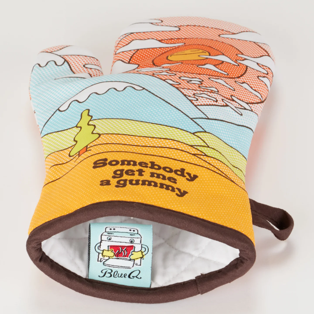 A novelty oven mitt with a colorful landscape print featuring mountains, a tree, and a sunset, with the text 'Somebody get me a gummy' on the front. The mitt has a white interior lining and a visible BlueCo brand tag.