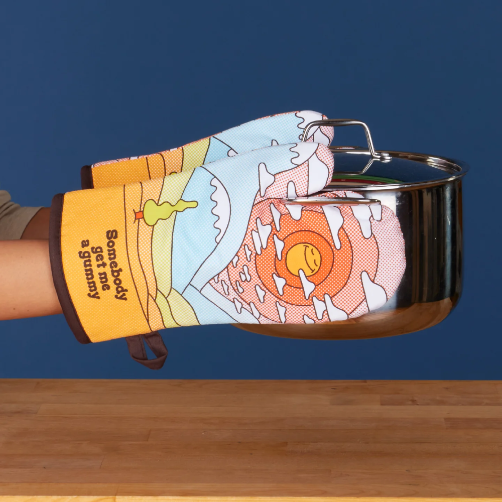 A person's hand wearing a colorful oven mitt with a cartoon landscape design and the text 'Everybody get me a gummi' holds a stainless steel pot over a wooden table against a dark blue background.