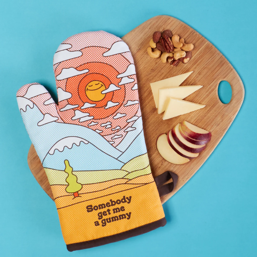 A novelty oven mitt with a colorful landscape illustration and the text 'Somebody get me a gummy' is styled on a bright blue background next to a wooden cutting board with cheese, apple slices, and nuts.