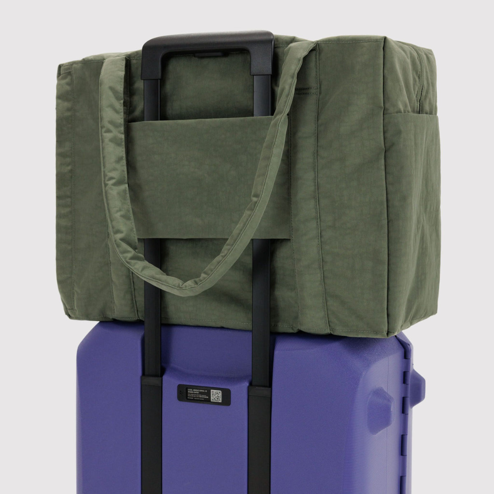 An olive green soft-sided duffel bag with a top handle and shoulder strap, shown resting on top of a purple rolling suitcase against a light gray background.