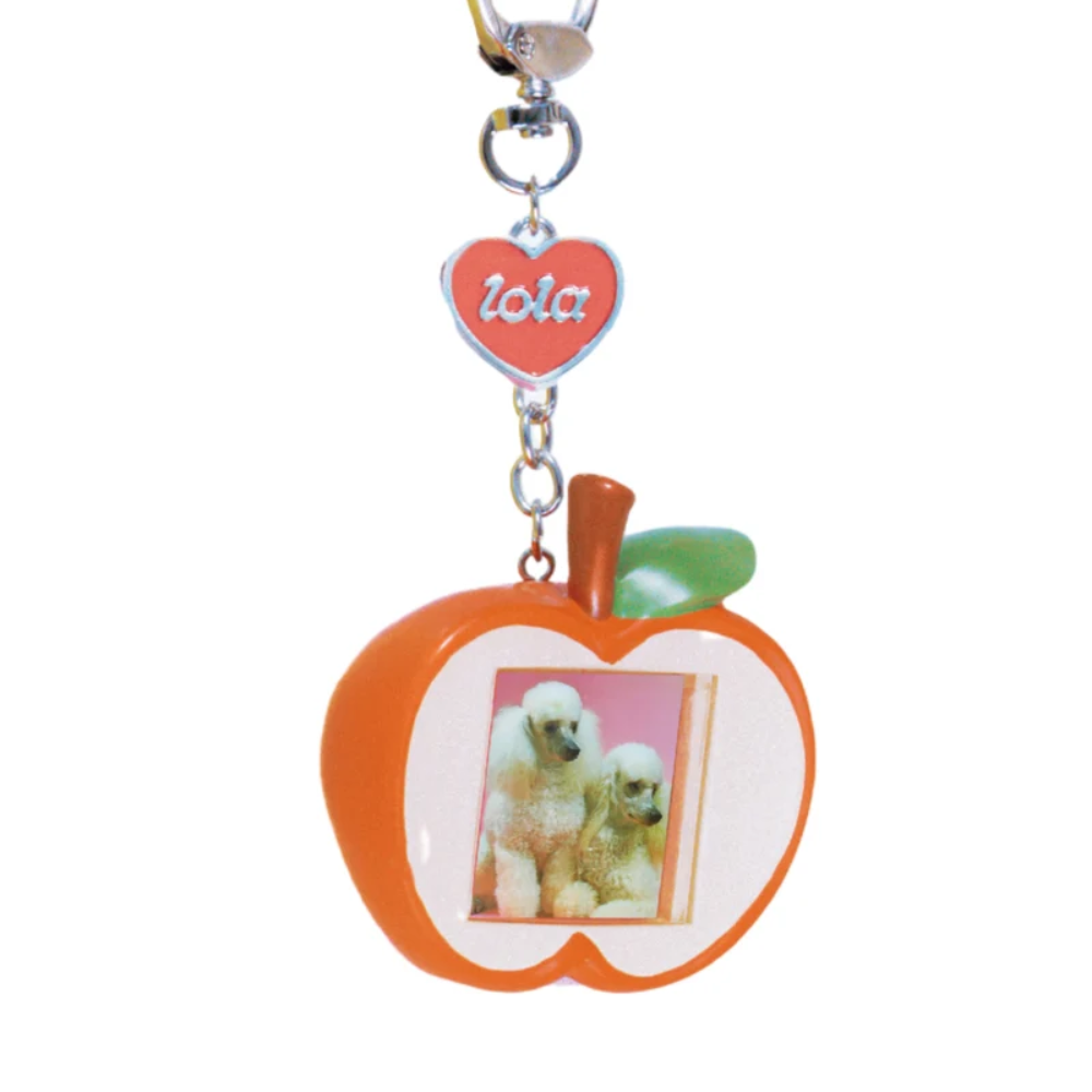 An apple-shaped keychain with a custom photo insert, featuring an orange body, green leaf, brown stem, a red heart charm with the name 'Iola' in white, and a silver metal key ring.