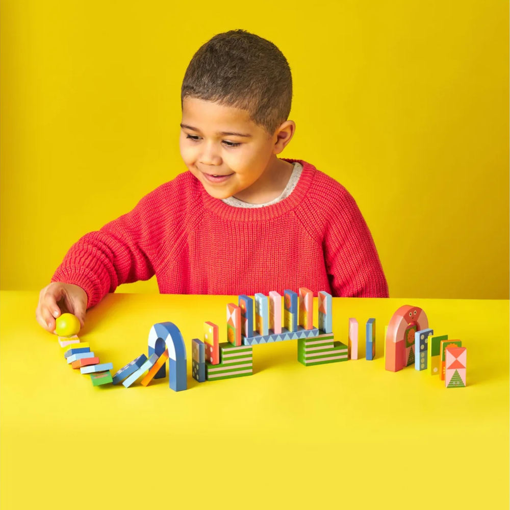 A young boy wearing a red knit sweater sits at a yellow table, playing with a colorful geometric wooden block domino game that has fallen in a short chain.