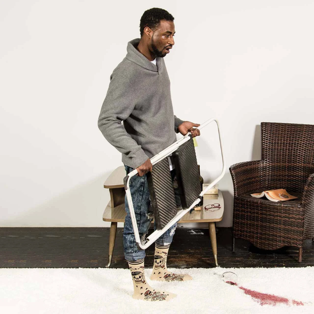 A man in a gray hoodie and patterned socks holds a white-framed mirror in a room with a wicker chair, side table, and white rug.