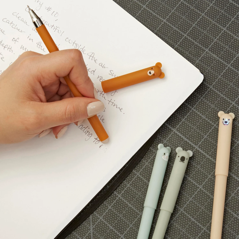 Hand holding a pen with a bear-shaped cap next to three other pens on a surface.