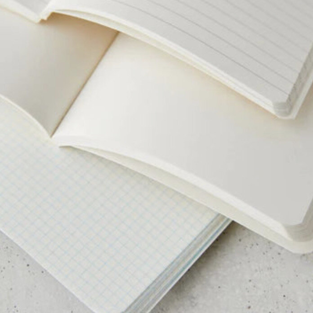 A close-up of stacked open notebooks showing lined pages on top and a light blue grid page underneath on a light textured surface.