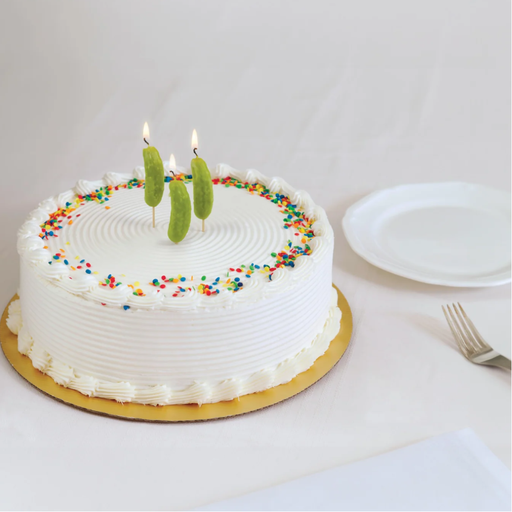 A round white frosted birthday cake on a gold cardboard base, decorated with colorful sprinkles and three green candle-shaped candles on top. A white plate and fork sit to the right on a white tablecloth background.