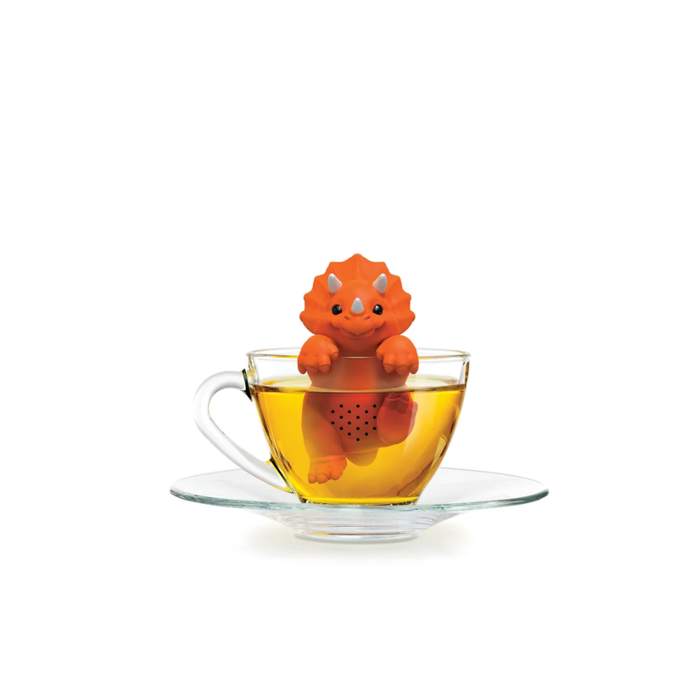 An orange dinosaur-shaped tea infuser sits inside a clear glass teacup filled with amber tea on a matching glass saucer, isolated on a white background.