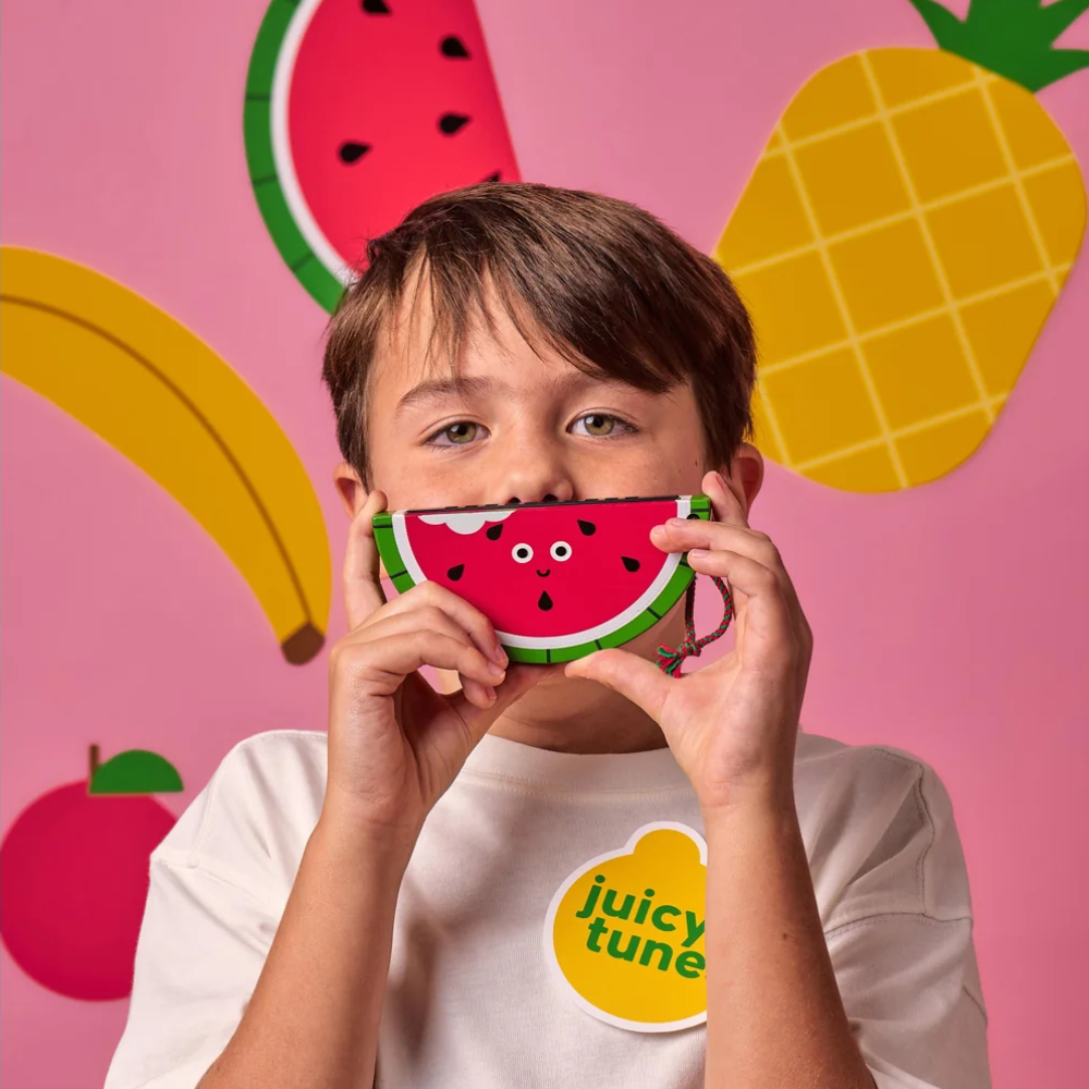 A young boy holding a watermelon-shaped harmonica in front of his face. The background is a pink wall decorated with paper cutouts of a pineapple, banana, watermelon, and apple.