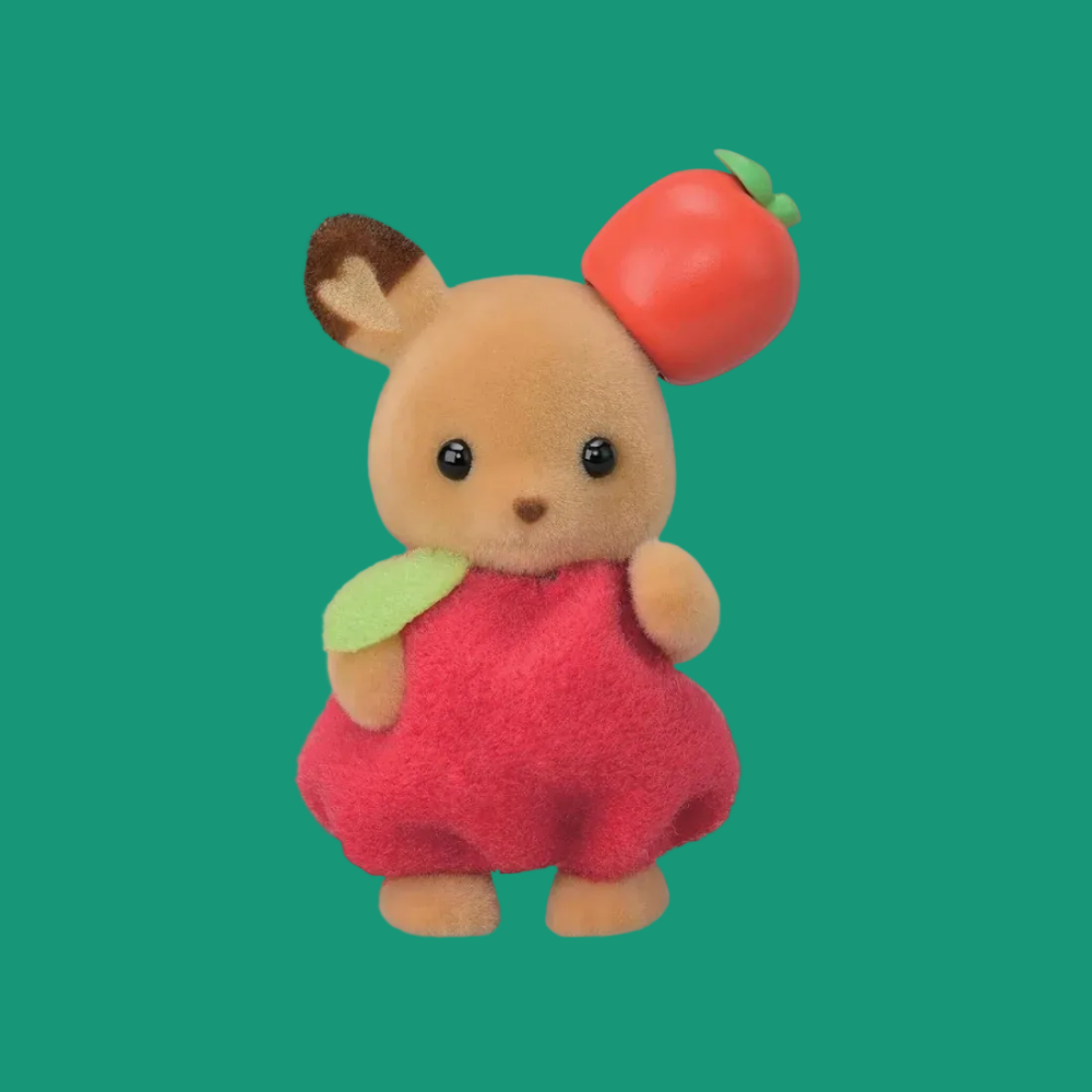 A small plush toy animal with tan fur, black eyes, and a bright red dress, holding a red apple with a green stem and leaf against a plain white background.
