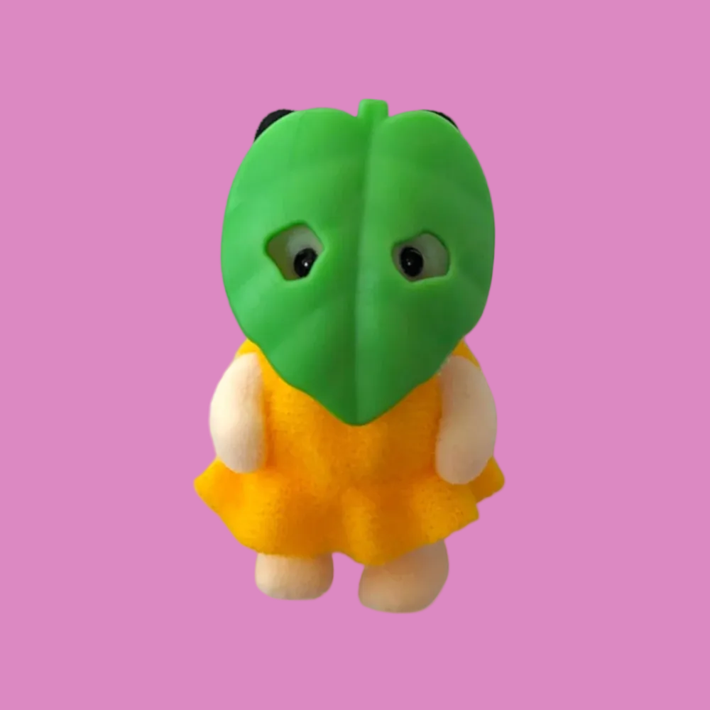 A small plush toy character with a green leaf-shaped head, black eyes, pale peach arms and legs, and a yellow-orange dress, shown on a plain white background.