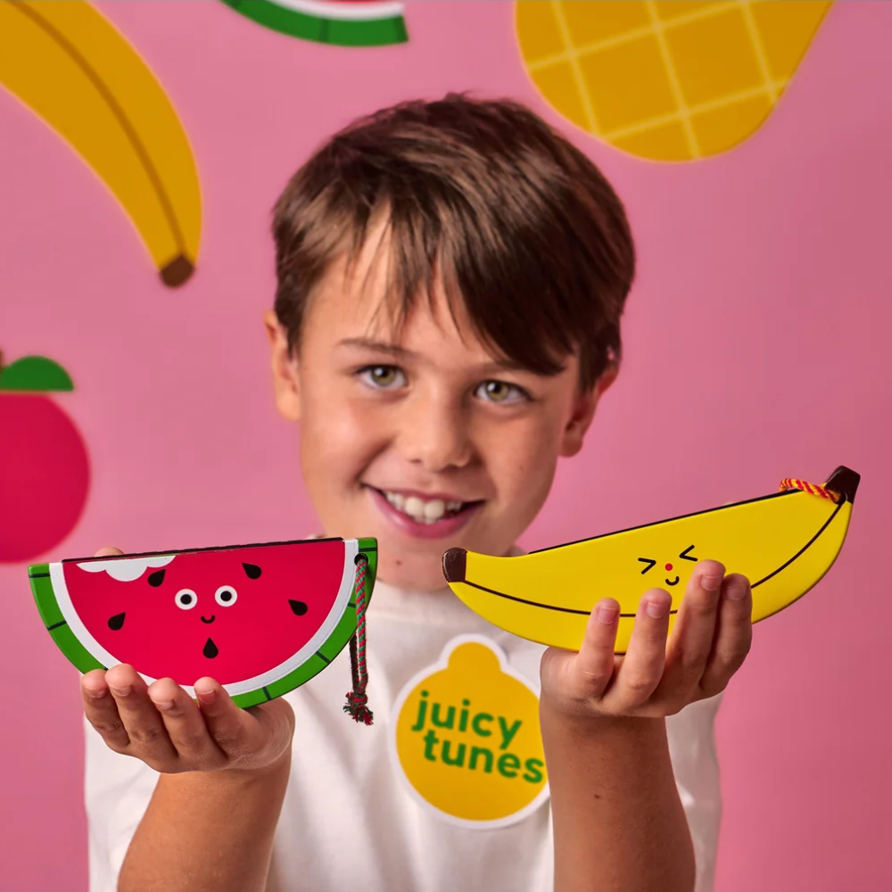 A smiling child holds two colorful fruit-shaped hamonicas, a watermelon slice and a banana, against a pink background with fruit illustrations. The child wears a white shirt with a yellow 'juicy tunes' logo.