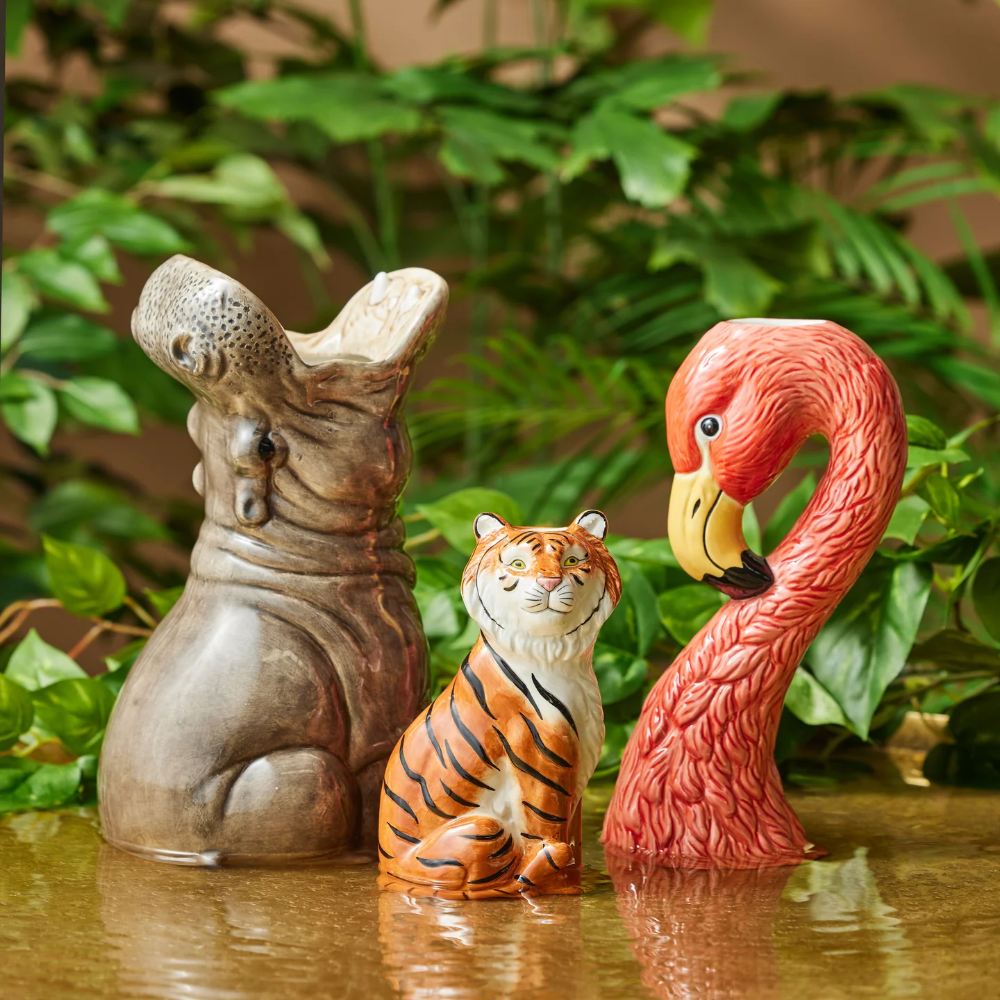Three animal figurines (hippo, tiger, flamingo) on a reflective surface with green foliage in the background.