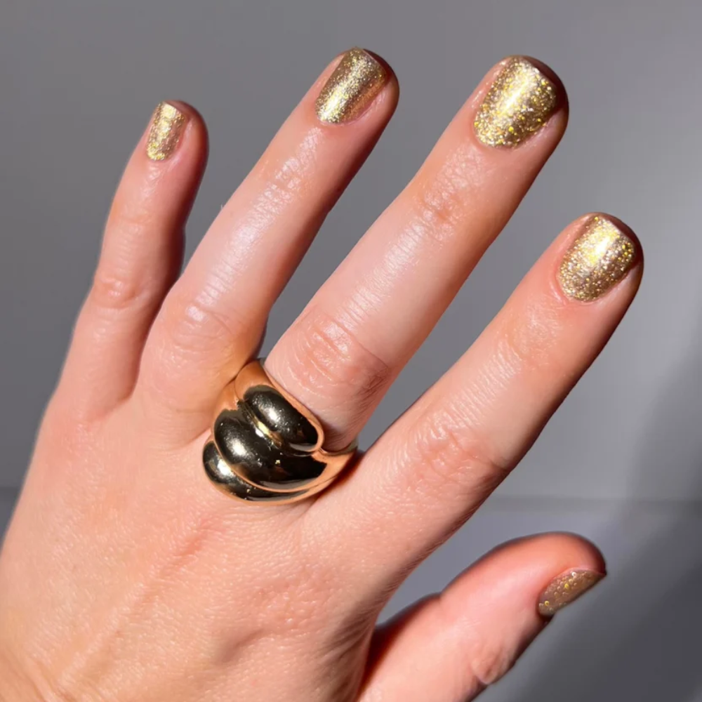 A close-up of a hand with gold glitter nail polish, wearing a chunky gold ring with a textured, ridged design. The background is a clean, neutral gray studio backdrop.