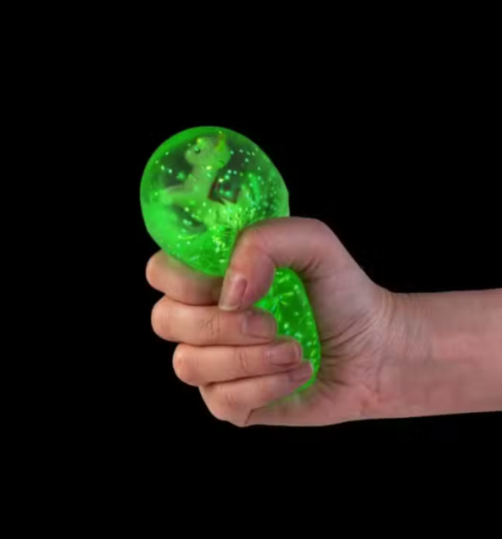 A hand squeezing a bright green, translucent stress ball toy with glitter and a small dinosaur figure inside, set against a solid black background.