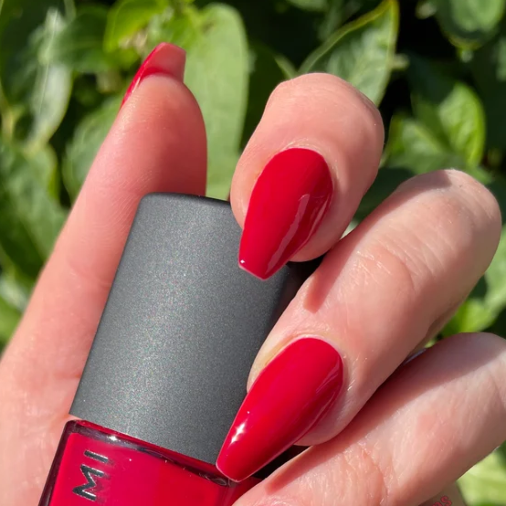 Hand holding a red nail polish bottle with green foliage in the background