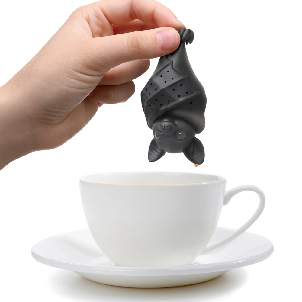 Hand holding a bat-shaped tea infuser over a white cup on a saucer.
