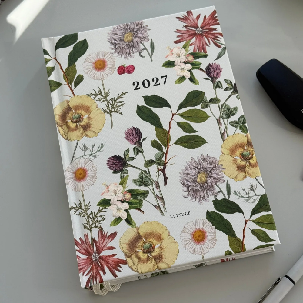 A 2027 floral planner or desk calendar lies open on a light gray surface, showing a botanical cover design with yellow, pink, purple, and green flowers and leaves. A pen and a dark object are partially visible around it.