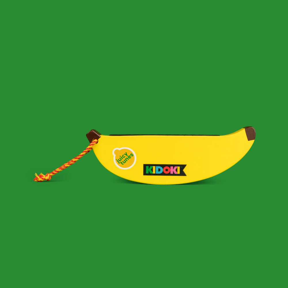 A yellow banana-shaped harmonica pouch with a braided orange rope handle, featuring the KIDOKI logo and a circular 'kidz hands' sticker, set against a solid green background.