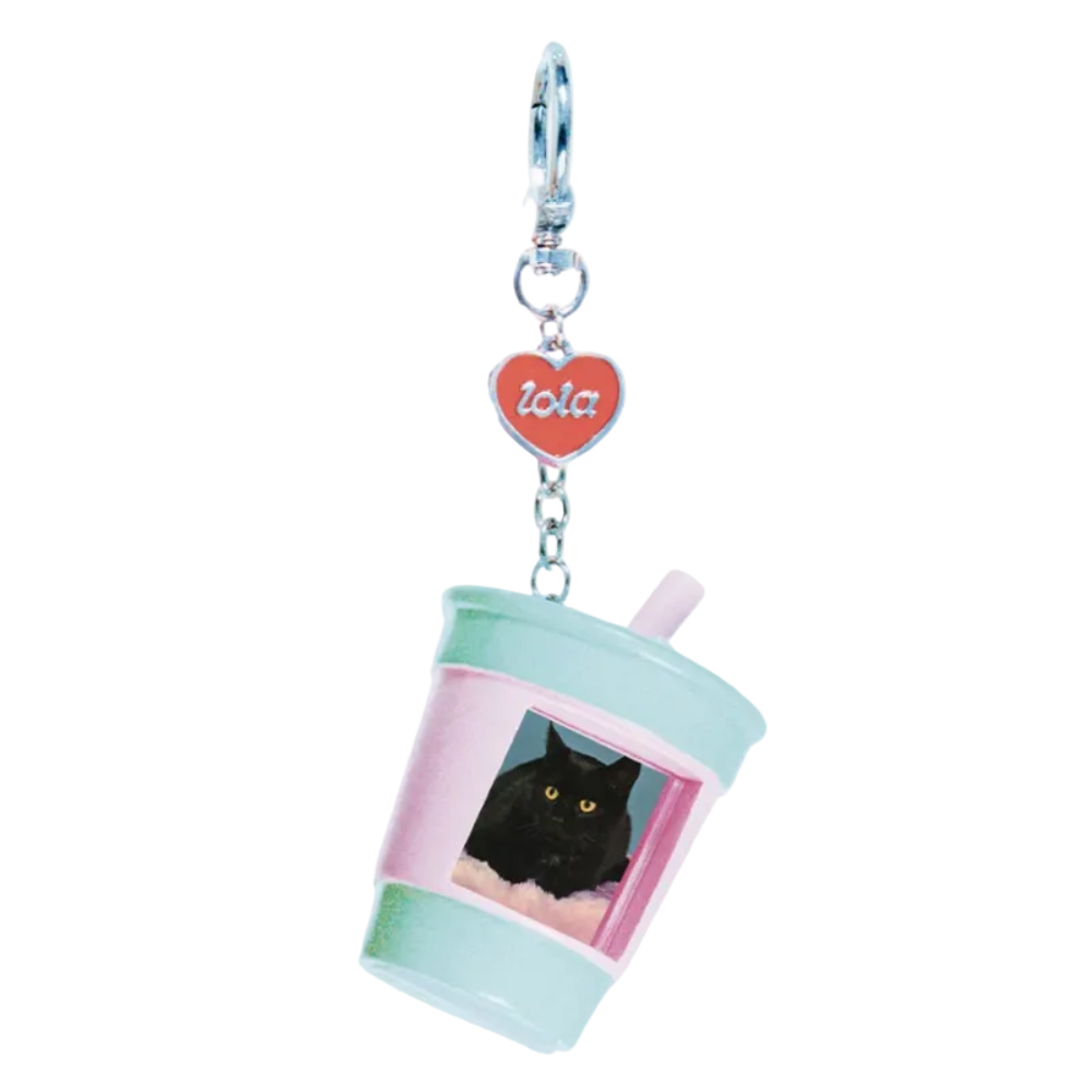 A novelty keychain charm shaped like a pastel drink cup with a black cat photo printed on the front, attached to a silver clasp and a red heart charm with the name 'lola' in white script, shown on a plain white background.