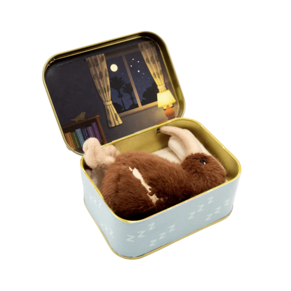 A brown plush sleeping rabbit toy nestled inside an open light blue tin with a gold rim. The tin lid features a printed illustration of a cozy nightstand and window at night.