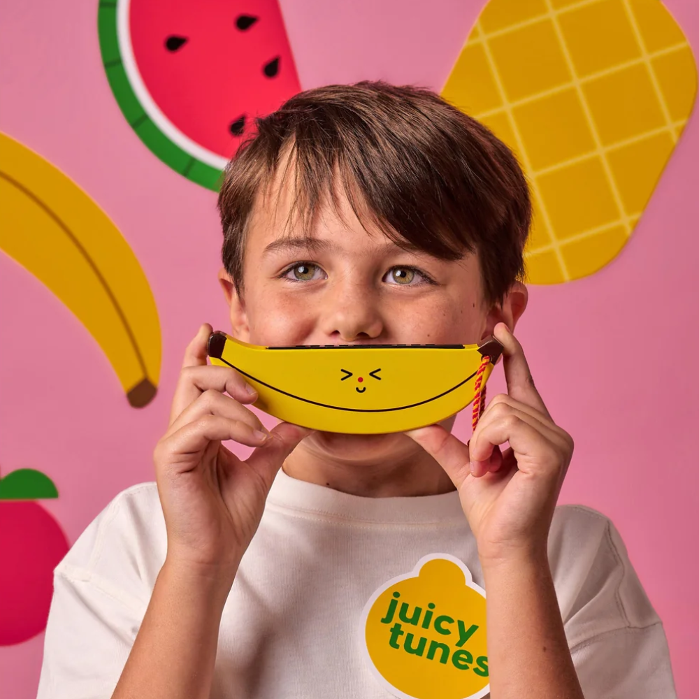 A young boy holding a yellow banana-shaped harmonica in front of his face. He is posing against a pink background decorated with paper cutouts of a watermelon, pineapple, and bananas.