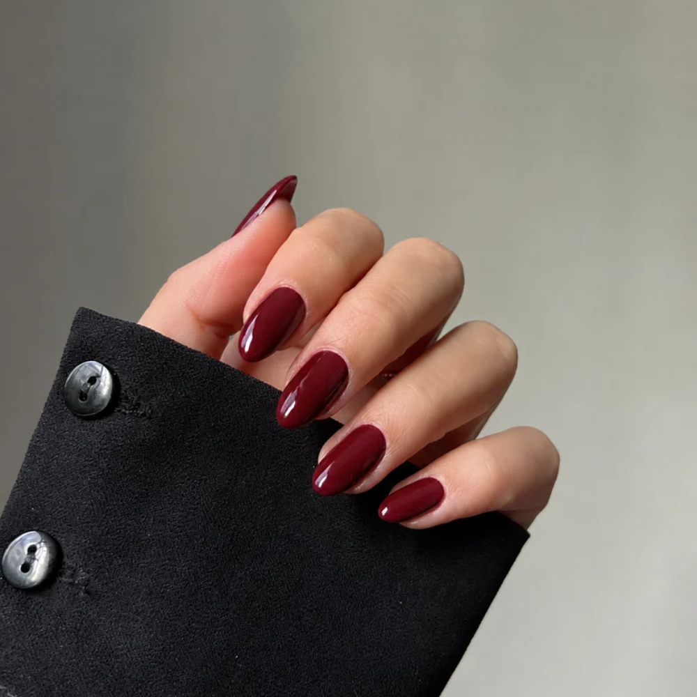 Hand with maroon nail polish wearing a black sleeve against a neutral background
