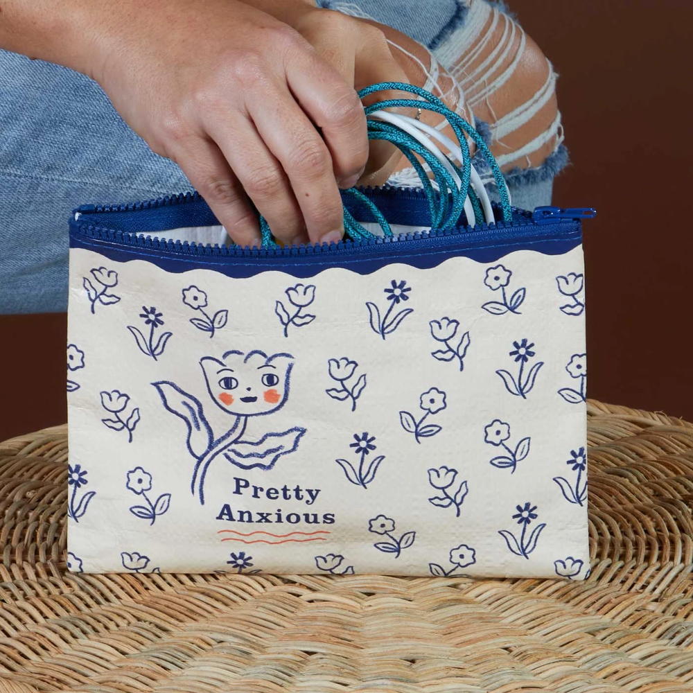 A cream canvas zip pouch with a blue pattern of flowers and the text 'Pretty Anxious' on the front, held by a person's hand. The pouch is resting on a woven wicker surface against a solid brown background.