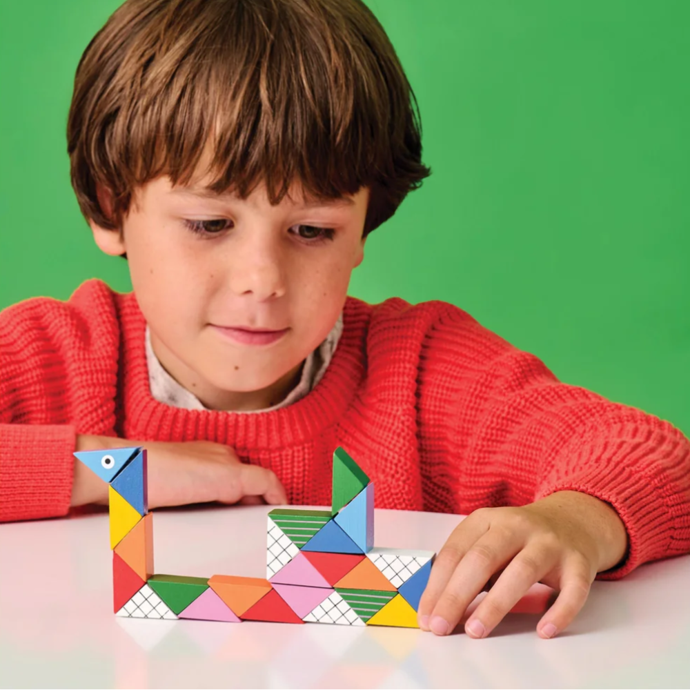 A young boy with brown hair wearing a red knit sweater sits at a white table, looking down at a colorful geometric toy he is building. The background is a solid green color.