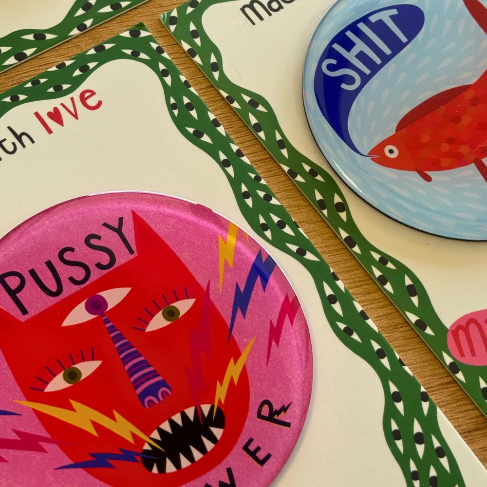 A close-up of printed cards or labels laid on a wooden surface, featuring bold text and colorful illustrations. The most prominent visible item is a pink circular label with a stylized red face and the word 'PUSSY' in black, bordered by green patterned lines.
