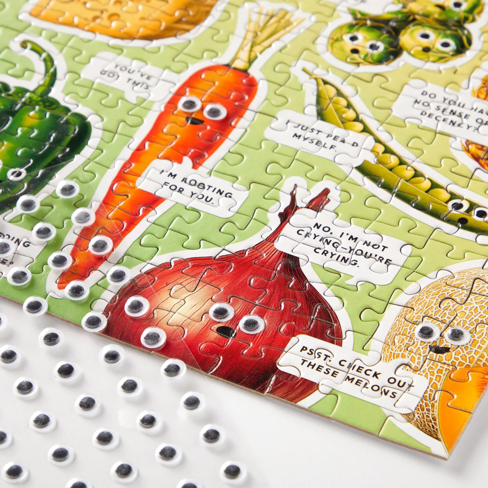 A close-up of a completed jigsaw puzzle featuring cartoon vegetables with funny faces on a light green background, with loose plastic googly eyes scattered on a white surface.