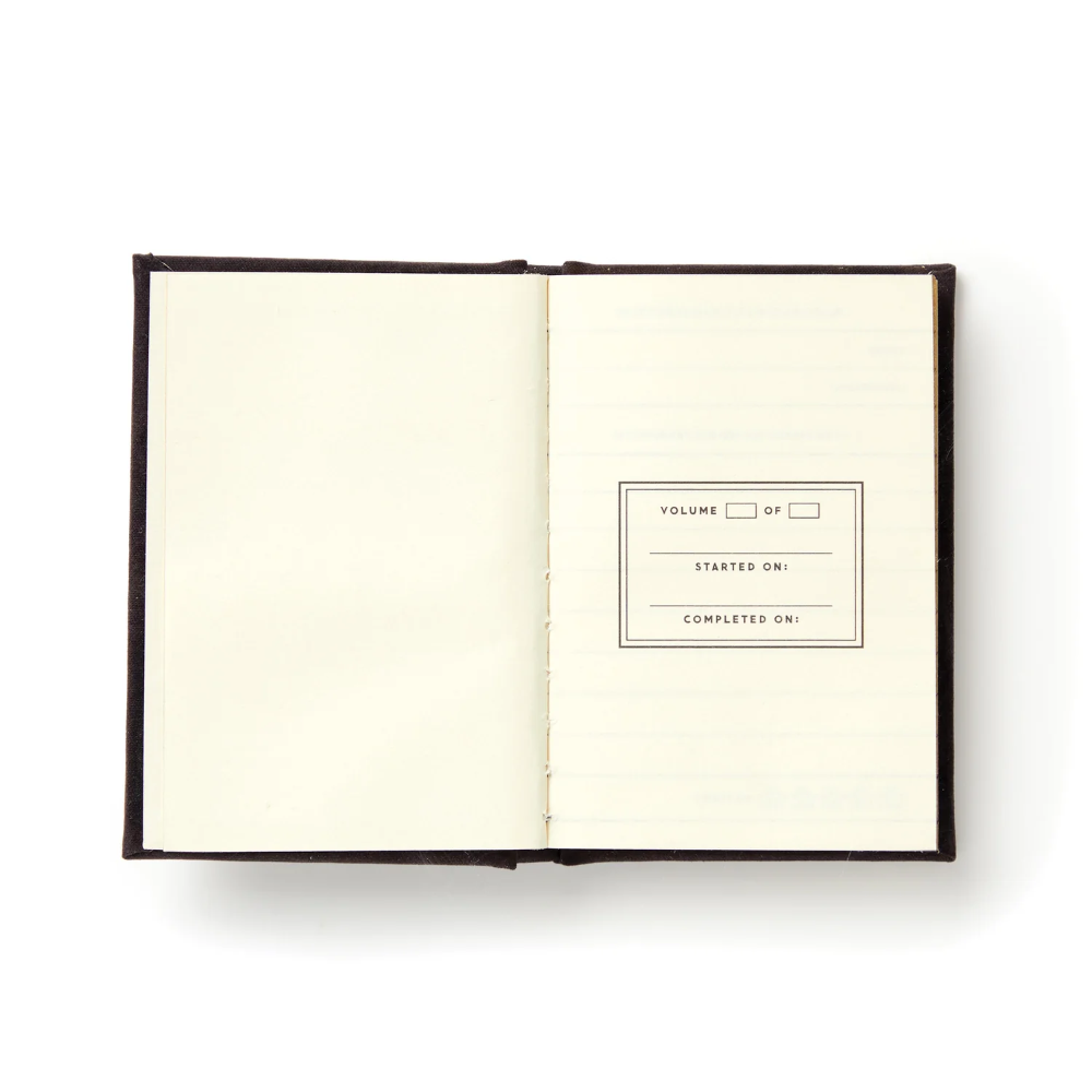 An open hardcover notebook with cream-colored lined pages, showing a dark brown cover and a printed label on the right page that reads 'VOLUME OF', 'STARTED ON:', and 'COMPLETED ON:'.