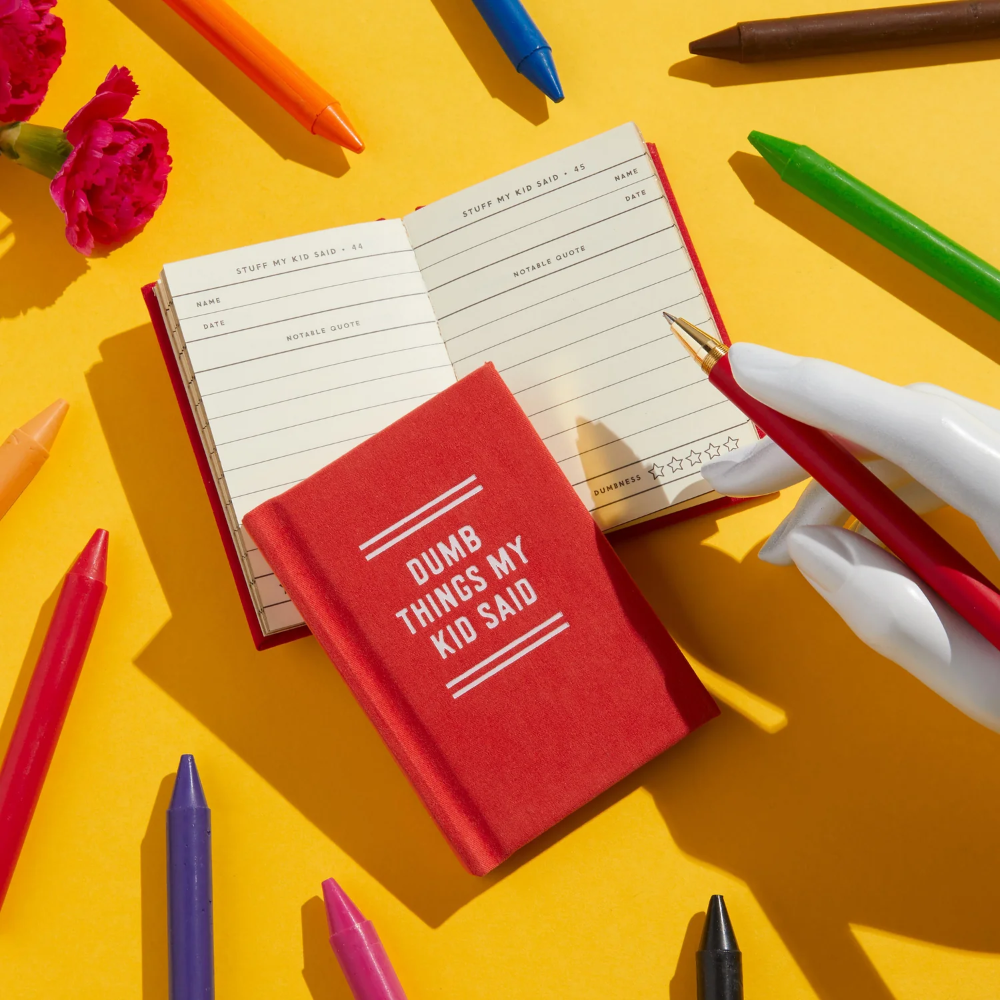 Open notebook with 'Dumb Things My Kid Said' on a red cover, surrounded by colorful pens and flowers on a yellow background.