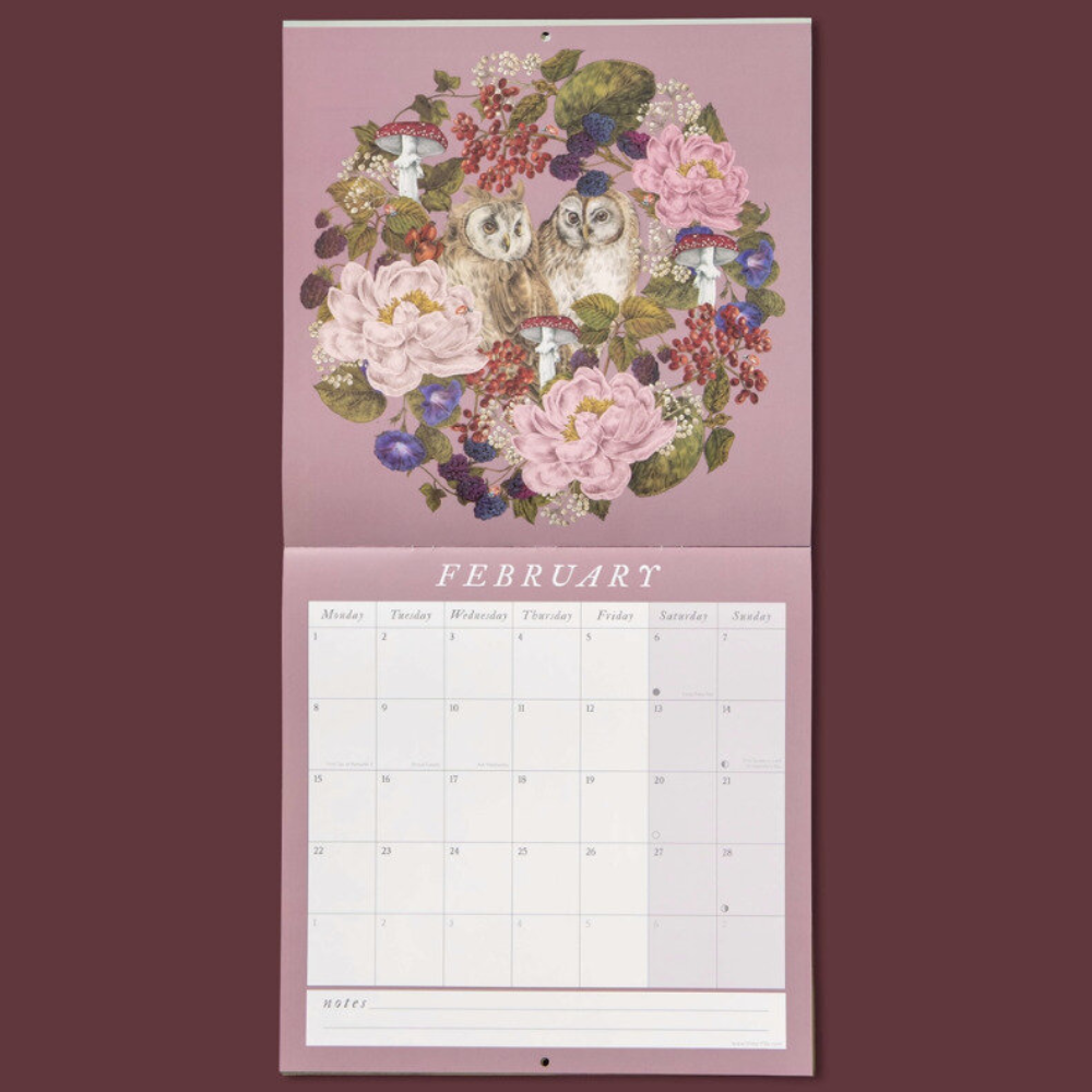 A hanging wall calendar page for February with a pink cover featuring an owl surrounded by flowers and berries, above a printed calendar grid.