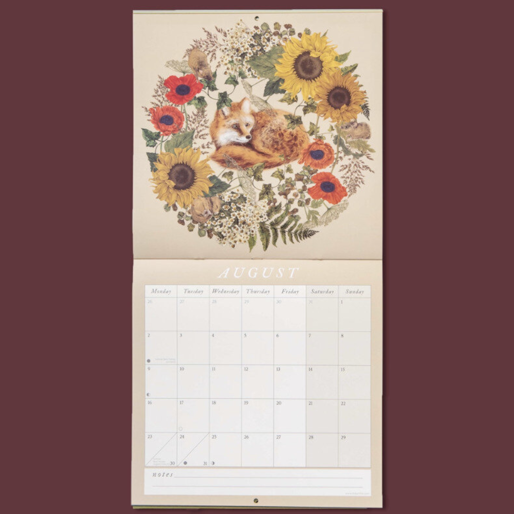 A hanging wall calendar page shown open against a dark burgundy background. The top page features an illustrated floral wreath with sunflowers, red flowers, and a curled-up fox at the center.