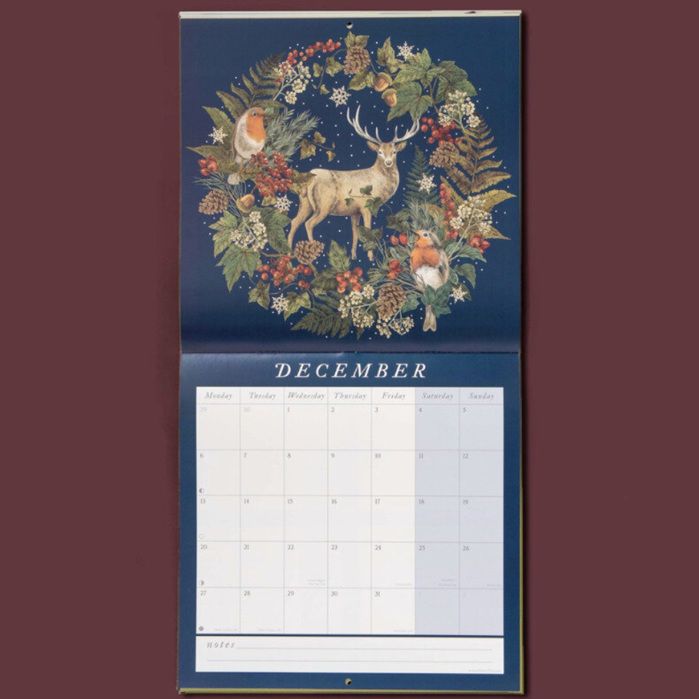 A hanging December calendar page with a dark blue background and an illustrated wreath featuring a stag, birds, berries, pinecones, and foliage. The lower section shows the monthly calendar grid labeled DECEMBER.