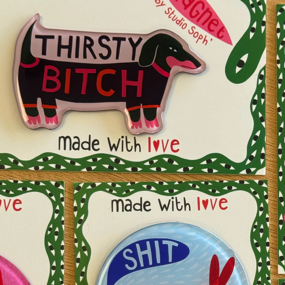 A close-up of enamel pins displayed on a decorated card. The main pin is shaped like a dachshund and reads 'THIRSTY BITCH' with a pink tongue and pink hooves.