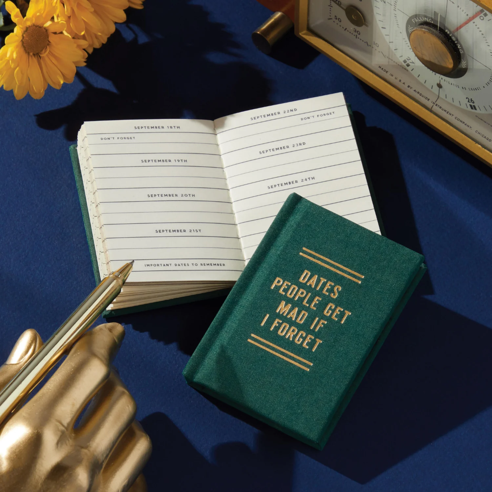 Small notebook with a humorous quote on a blue surface with a pen and flowers.