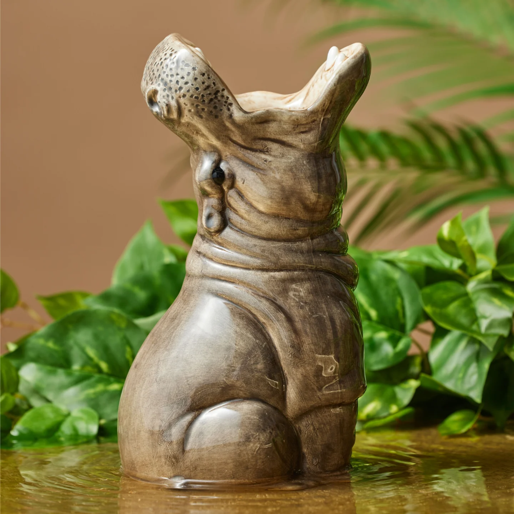A sculpted hippopotamus figurine with an open mouth, standing in shallow water with green foliage and a warm brown background.
