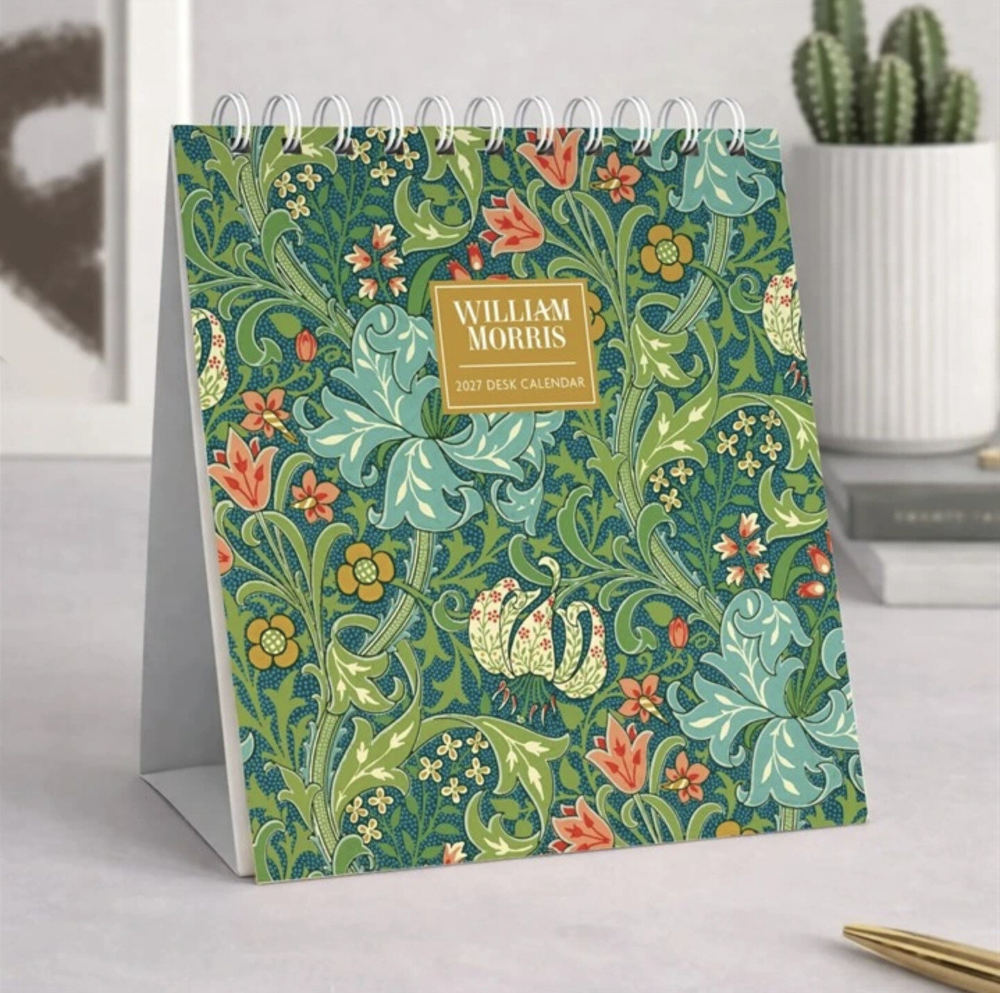 William Morris 2027 desk calendar standing on its own, featuring a detailed green and blue floral pattern with orange flowers and a gold label, set against a styled desk background with books and a cactus.