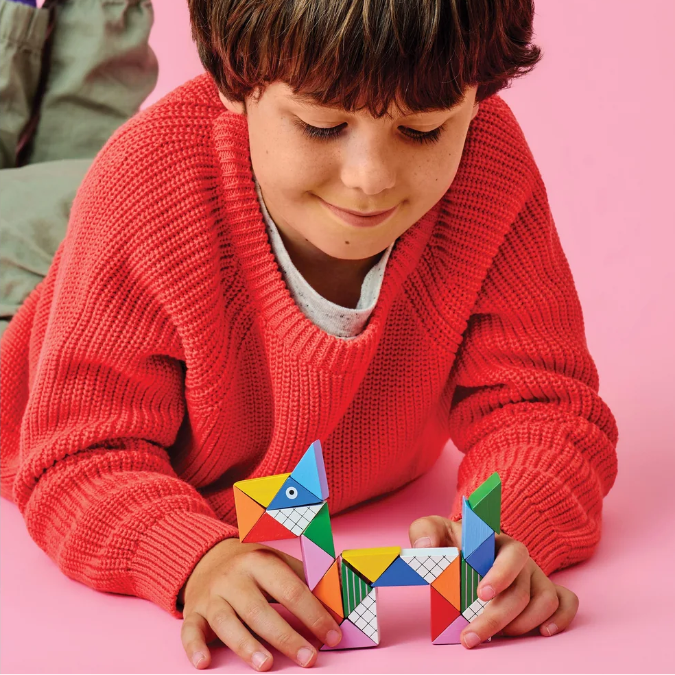 A young boy with brown hair lies on a pink floor, wearing a red knit sweater and purple socks while playing with a colorful geometric toy made of triangle pieces.