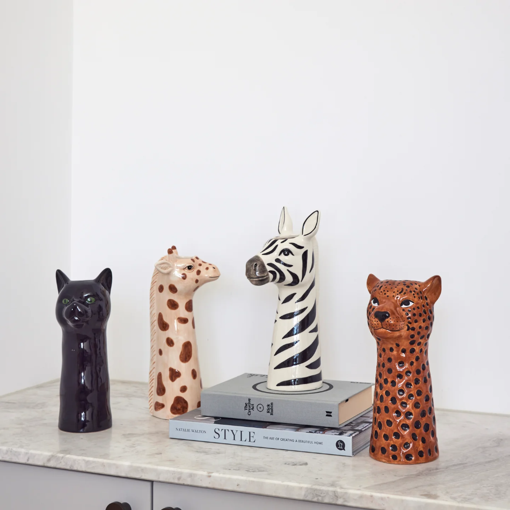 Four animal head ceramic figurines displayed on a marble shelf against a white wall: a black cat, a tan giraffe, a zebra, and a brown leopard, with books stacked underneath the zebra.
